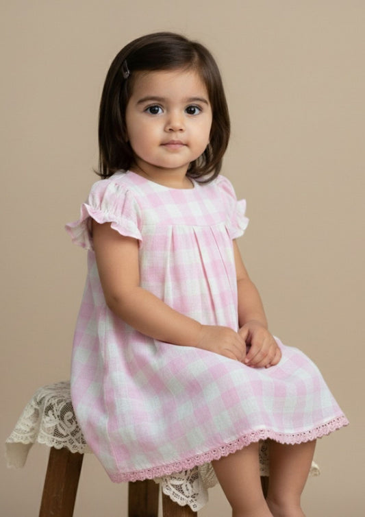 Baby girl sitting sideways wearing blush pink check cotton A-line dress with crochet lace