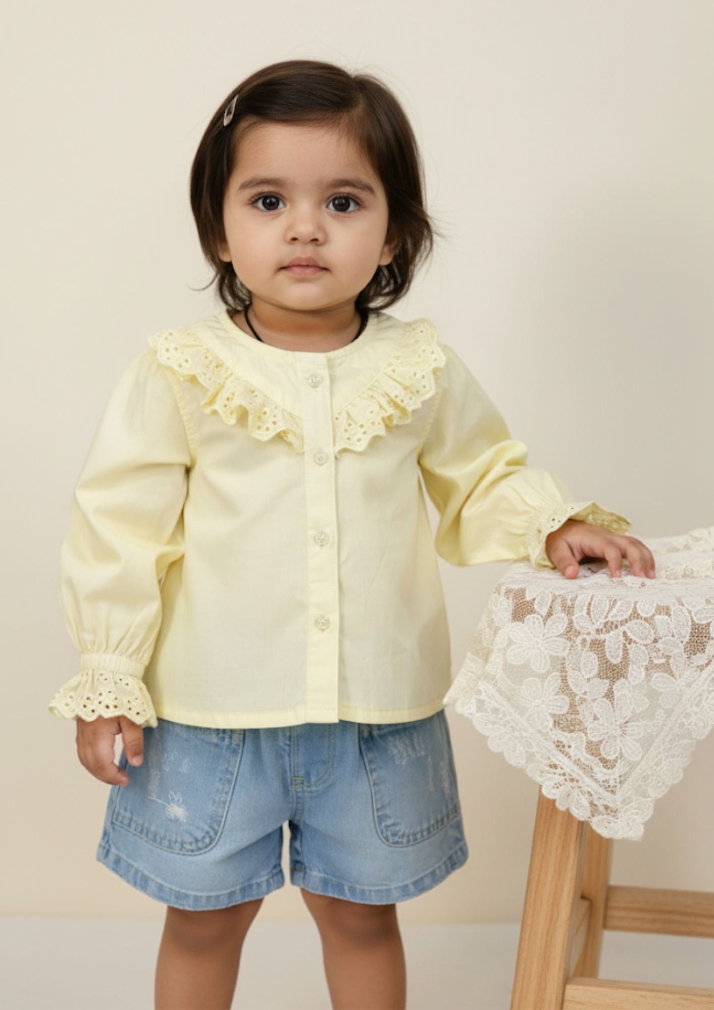 Baby girl standing with hand on table wearing buttery yellow cotton princess top and light blue denim shorts