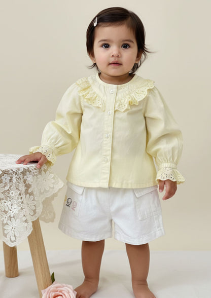 Baby girl standing with hand on table wearing buttery yellow cotton full sleeve princess top and white smiley shorts