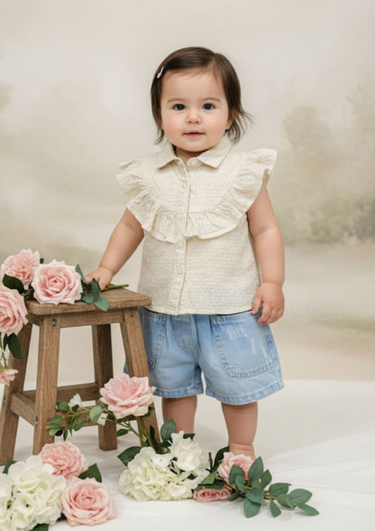 Baby girl standing with hand on stool wearing cream sleeveless accoba top and light blue denim torn shorts